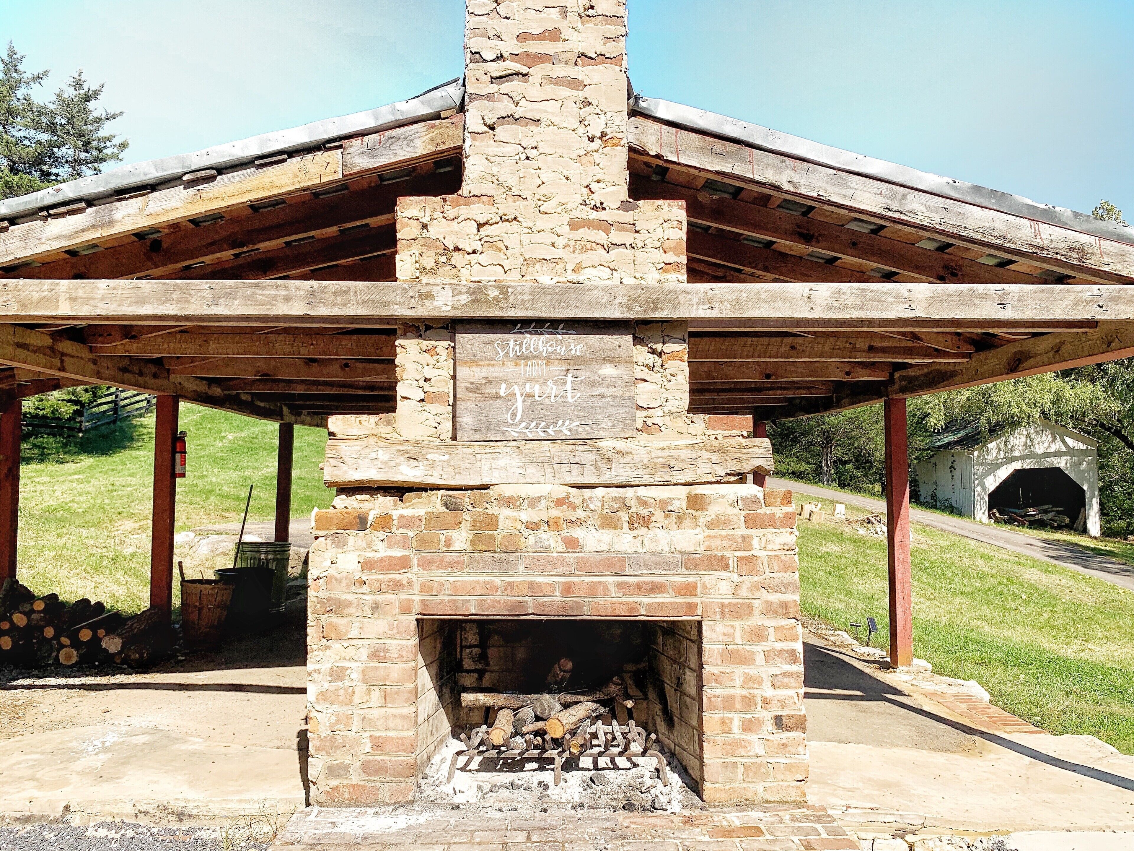 A rustic stone fireplace with a wooden roof structure, surrounded by a grassy area and a stone building in the background.