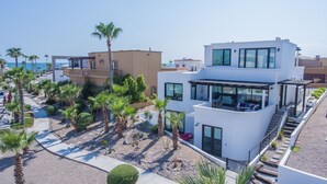 A modern multi-story building with a rooftop patio, surrounded by palm trees and a clear view of the sea. - Breathtaking Custom Home in Sandy Beach with Great Amenities! (Puerto Penasco)