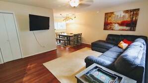 A living room with a sofa, a coffee table, a dining area with a table and chairs, a TV mounted on the wall, and a painting on the wall.