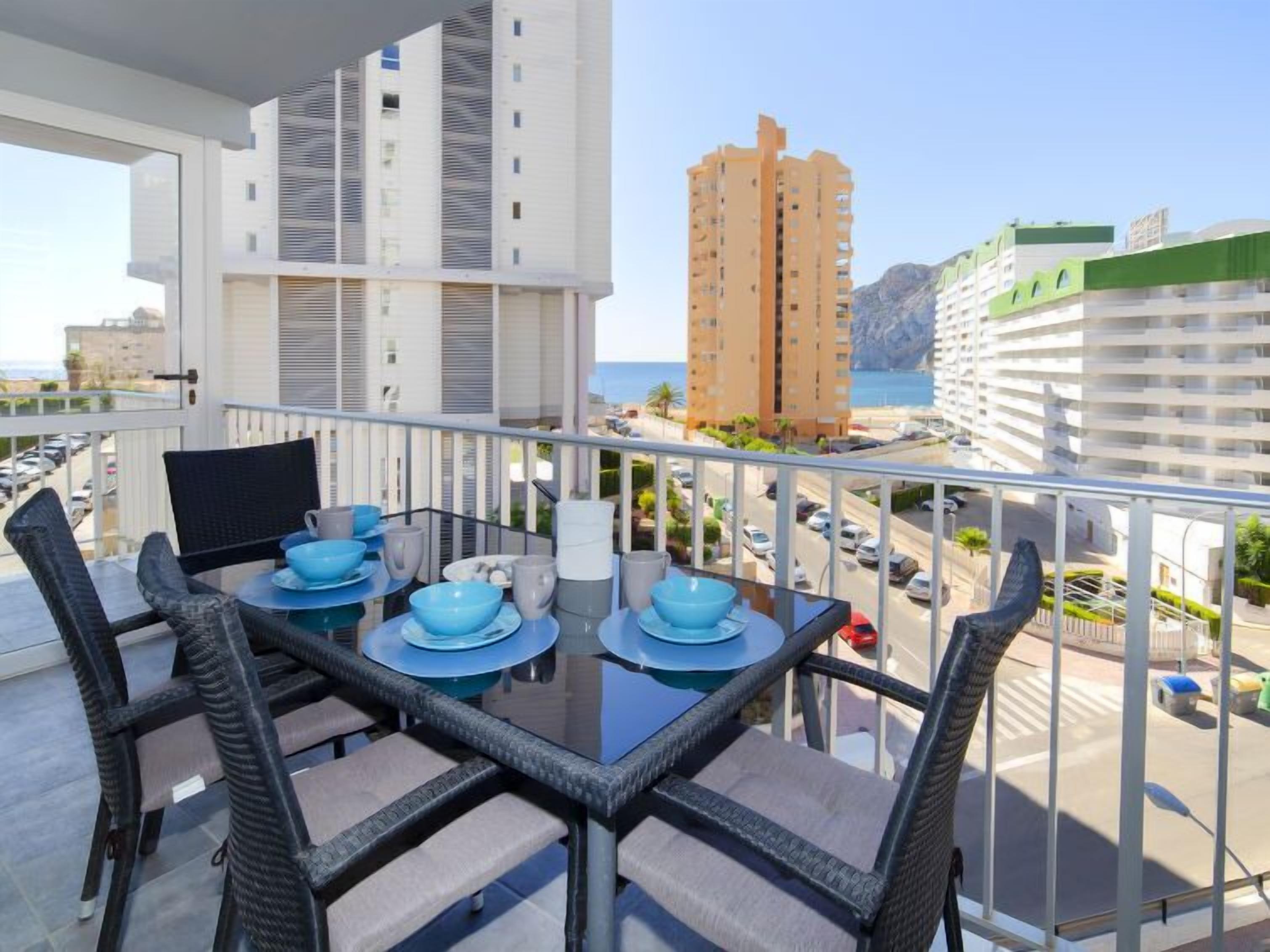 A balcony with a dining table set for four, overlooking a coastal cityscape with modern buildings and a view of the sea.