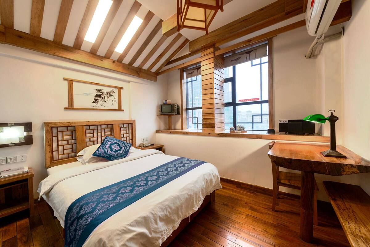 Ein Schlafzimmer mit Holz-Kopfteil, einem Bett mit blau kariertem Überwurf, einem Holztisch mit Lampe und einem Fenster mit Blick auf die Stadt.