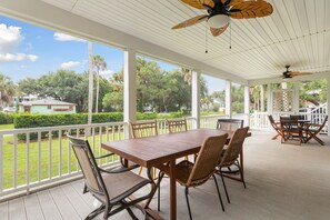 Eine überdachte Terrasse im Freien mit einem Holztisch und -stühlen, einem Deckenventilator und Blick auf die umliegende Natur und Häuser.