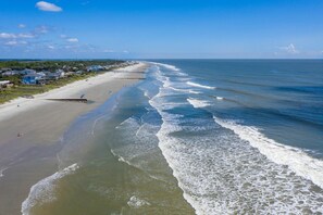 Ein Strand mit einer langen Küstenlinie, Häuser entlang des Strandes und ein Steg, der sich ins Wasser erstreckt.
