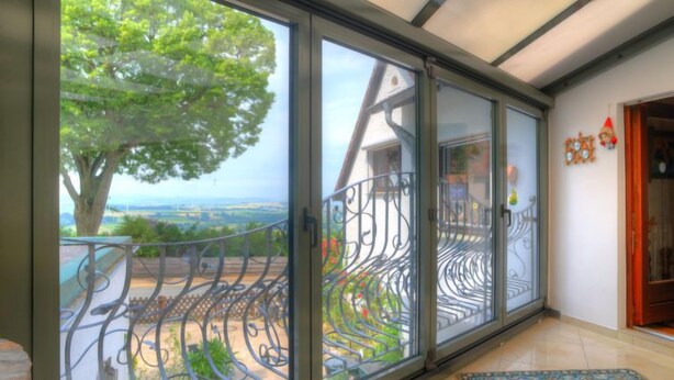 Une pièce dotée de grandes portes-fenêtres coulissantes donnant sur un balcon avec une rambarde en métal, d’où l’on peut admirer un arbre et un paysage, et dont le sol est recouvert d’un tapis à motifs.