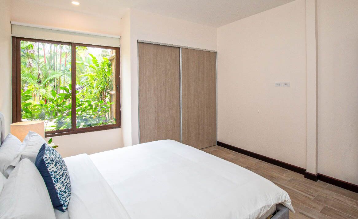 A bedroom with a large bed, a window with a view of greenery, and a sliding door closet.