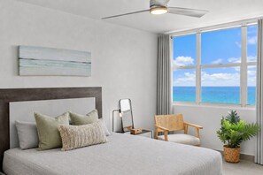 Un dormitorio con cama, una silla, una mesita de noche, una lámpara, un ventilador de techo, una ventana con vista al océano y un cuadro en la pared.