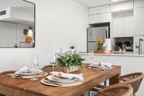 Una cocina moderna con armarios blancos, un refrigerador de acero inoxidable y un comedor con una mesa de madera dispuesta para dos.