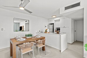 Une cuisine moderne avec des meubles blancs, un îlot central et un coin repas comprenant une table en bois et des chaises en osier.
