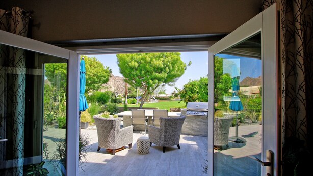 Une porte-fenêtre coulissante donne sur une terrasse extérieure aménagée avec des meubles en osier, un barbecue et offre une vue sur un jardin paysager agrémenté d’arbres et, au loin, sur une montagne.