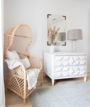 Een rieten stoel met kussens, een witte dressoir met lades, een spiegel en een lamp.