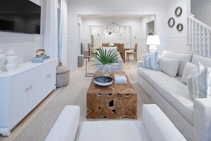 A modern living room with a white sofa, a wooden coffee table, and a dining area in the background.