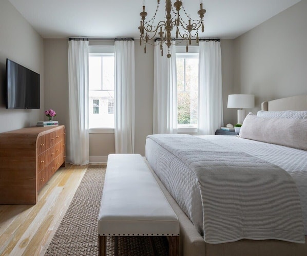 A bedroom with a large bed, a wooden dresser, a bench, a chandelier, and a TV on the wall.