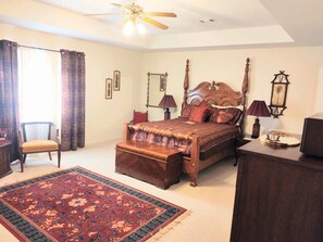A four-poster bed with a wooden headboard, a red and blue patterned area rug, a wooden nightstand, a chair, and a desk with a lamp. - Welcome to Polly's Place (Rockwell)