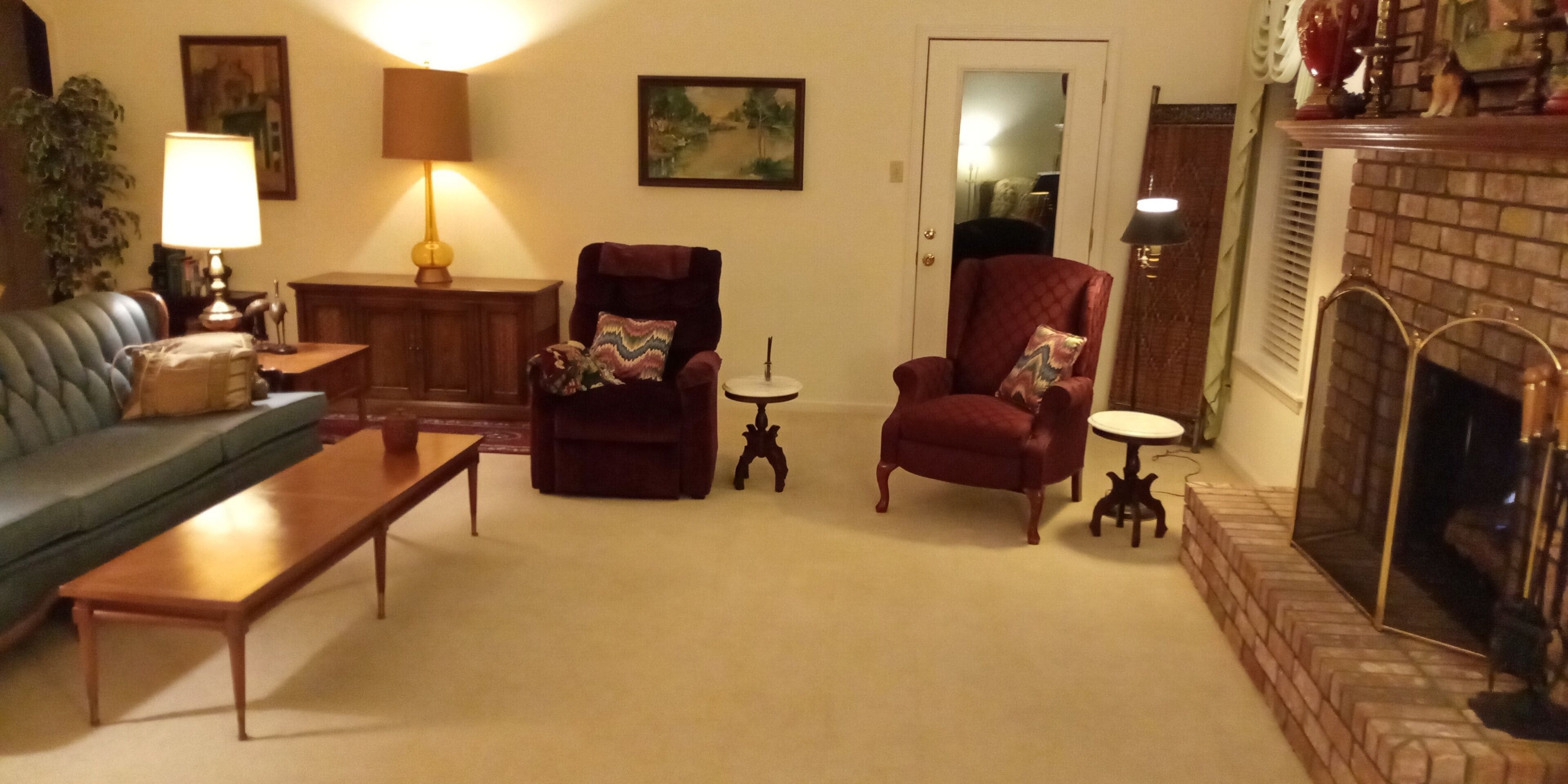 Uma sala de estar com um sofá verde, uma mesa de centro de madeira, duas poltronas com almofadas estampadas, uma mesa redonda pequena, um abajur, um quadro e uma lareira.
