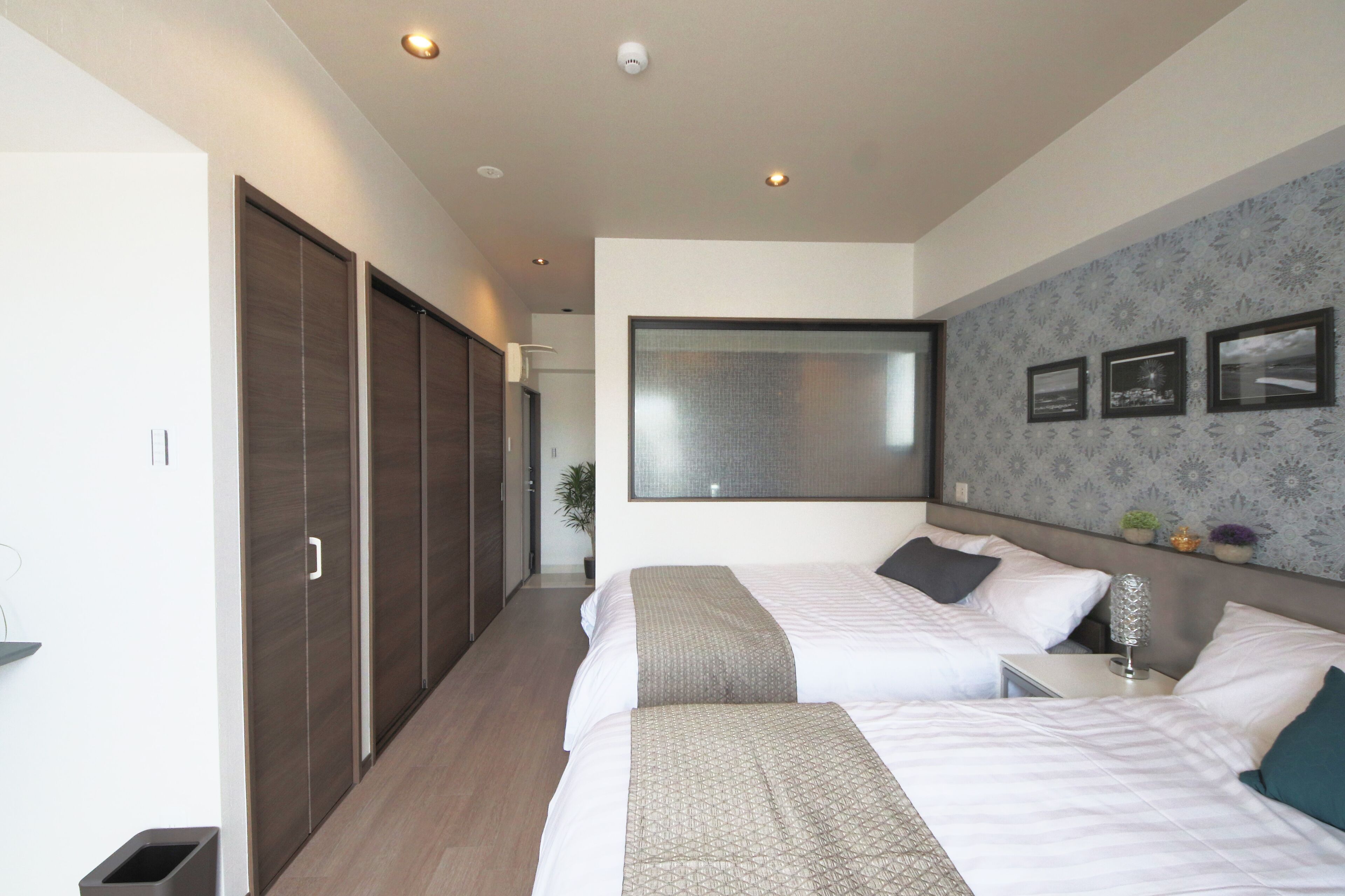 Habitación de hotel con dos camas, un ventanal grande, un armario y una mesita con lámpara.