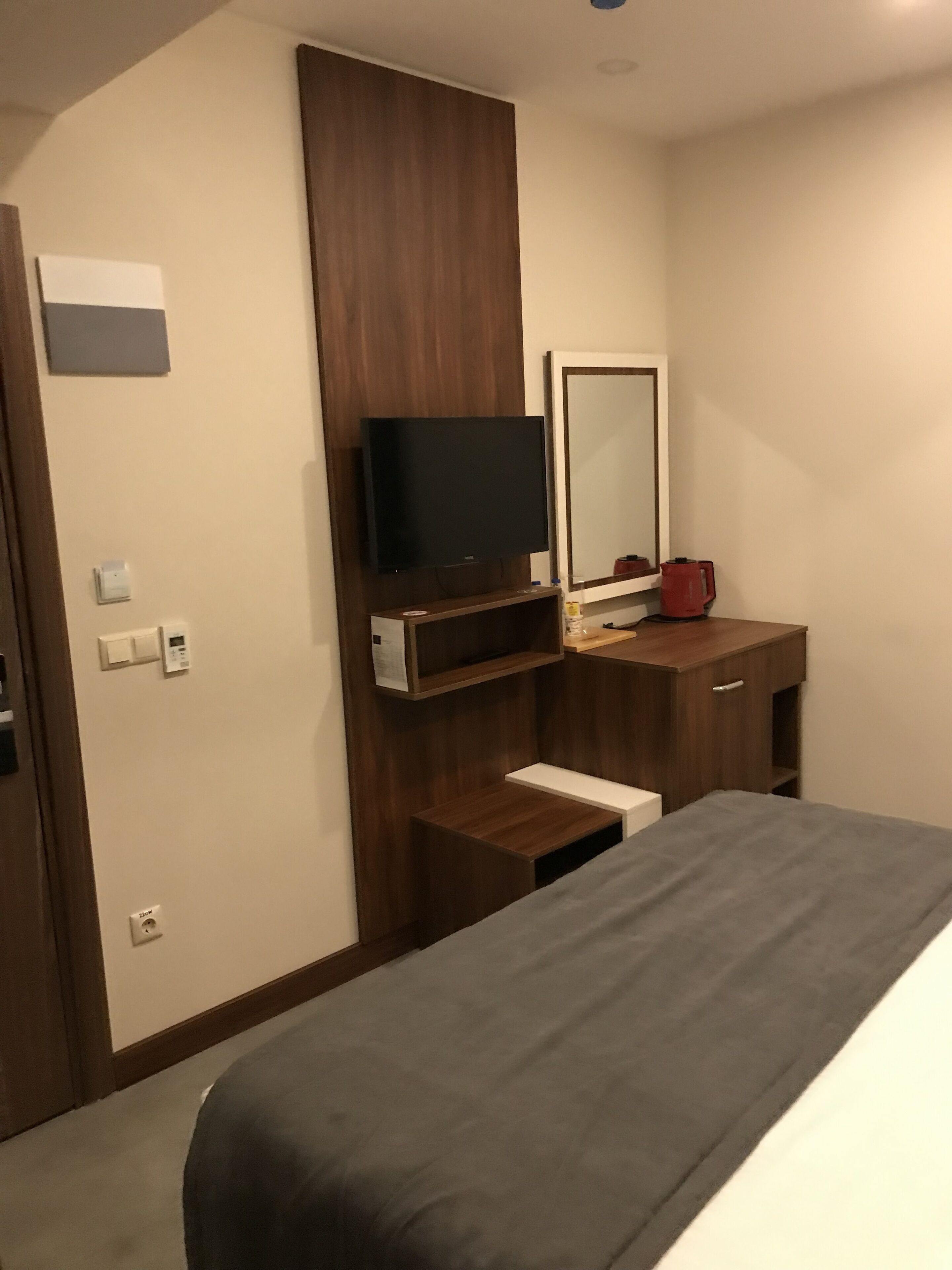 Una camera d'albergo con un letto, una TV fissata a un pannello di legno, uno specchio, un bollitore rosso e un piccolo ripiano.