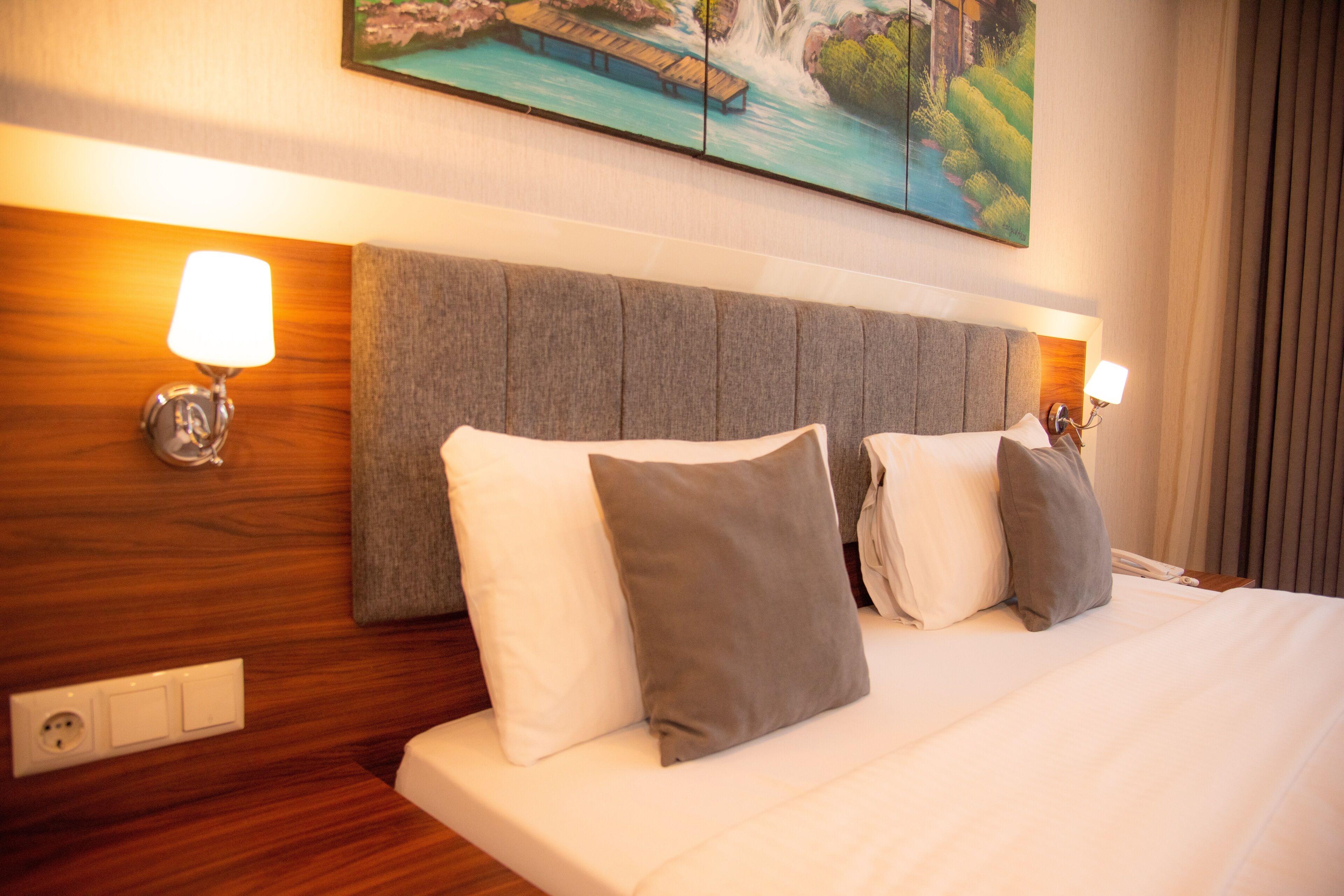 Habitación de hotel con una cama grande, dos lámparas de noche, cabecera de madera y un cuadro sobre la cama.