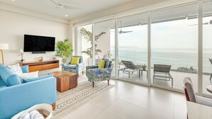 Una sala de estar moderna con un televisor grande, asientos cómodos, una mesa de centro y vistas al océano a través de amplios ventanales.