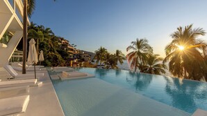 Zona junto a la piscina con sillones de descanso y sombrillas, rodeada de palmeras y edificios.