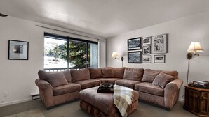 A living room with a brown sectional sofa, a small ottoman, a coffee table, and a sideboard. There are framed pictures on the wall, a floor lamp, and a window with a view of trees and mountains. - Great Weekend Destination-Between Snowbird & Alta, Hot Tub, BBQ, Free Parking, Free Ski Rental (Alta)