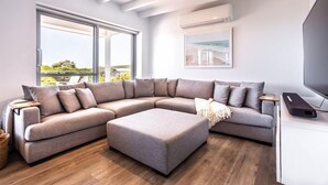A modern living room with a large L-shaped sectional sofa, a small ottoman, a flat-screen TV, and a large window offering a view of greenery. - Coastal home in Gracetown - relaxing! (Gracetown)