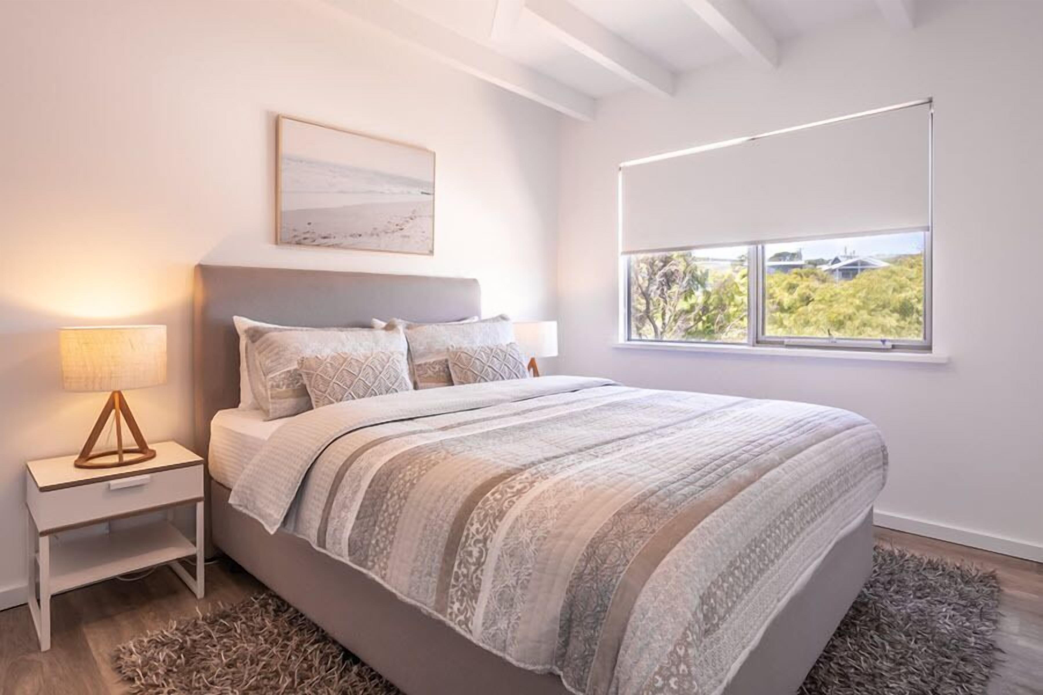 A bedroom with a bed, bedside lamps, a window with a view, and a painting on the wall.