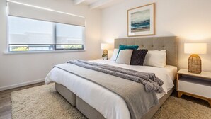 A modern bedroom with a large bed, a window with blinds, a bedside table with a lamp, and a framed picture on the wall. - Coastal home in Gracetown - relaxing! (Gracetown)