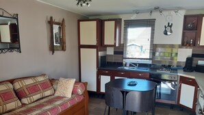 A compact living space with a sofa, a coffee table, a dining table, and a kitchen area including a stove and sink. - Chalet 100 m from the little sea of Gâvres (Riantec)