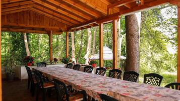 Uma área de refeições coberta ao ar livre, com uma mesa comprida e cadeiras, rodeada por árvores.