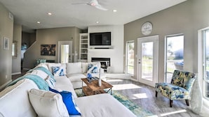 A spacious living room with a large sectional sofa, a coffee table, and a patterned armchair. A flat-screen TV is mounted above a fireplace, and there are large windows providing natural light. - Ocean-View 'Kahala Beach Bliss' Home in Galveston! (Galveston)