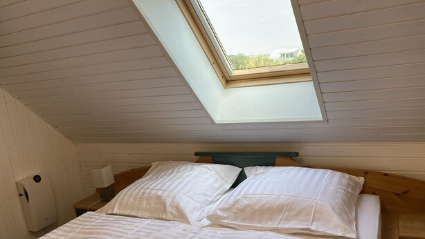 Une chambre avec un plafond en pente et un puits de lumière, comprenant un lit avec une literie blanche et une tête de lit en bois.
