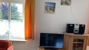 Un salon avec un meuble TV en bois, un téléviseur à écran plat, deux haut-parleurs, un fauteuil rouge, une fenêtre avec des rideaux et un tableau accroché au mur.
