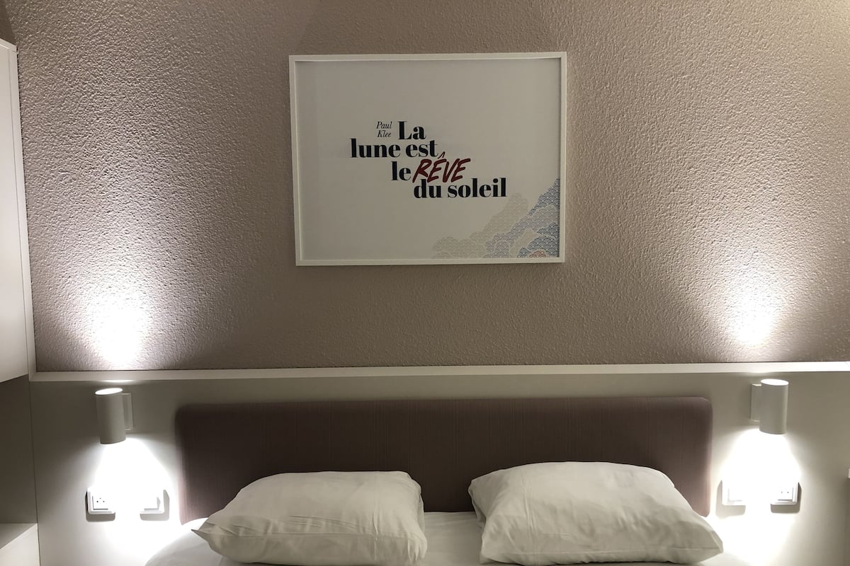 A hotel room with a bed, two pillows, a headboard, and a framed quote on the wall.