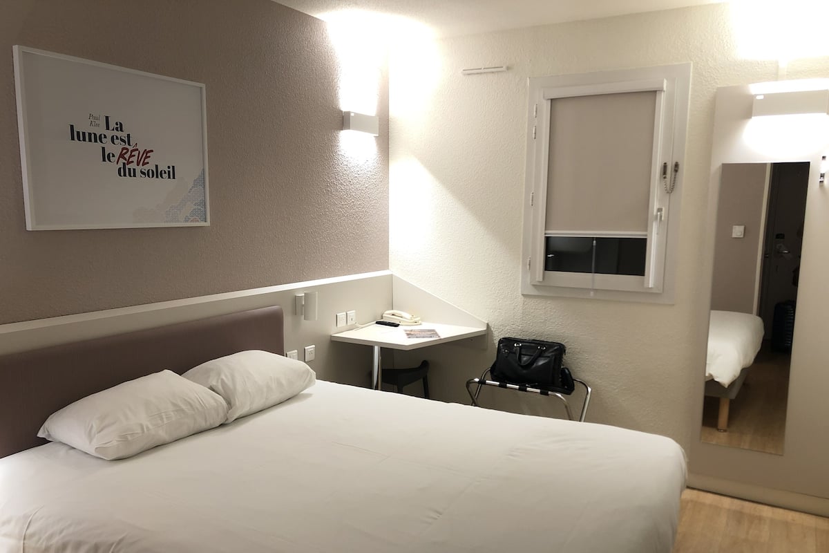 A hotel room with a bed, bedside table, lamp, and a small desk with a chair.
