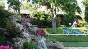 Ein Swimmingpool mit Wasserfall, umgeben von einem Zaun, und im Hintergrund ein Kinderspielbereich.