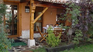 A rustic wooden building with a stone-paved entrance, a wooden table with chairs, and a garden area with various plants. - Basic Triple Room: Private entrance and porch (Olmedo)