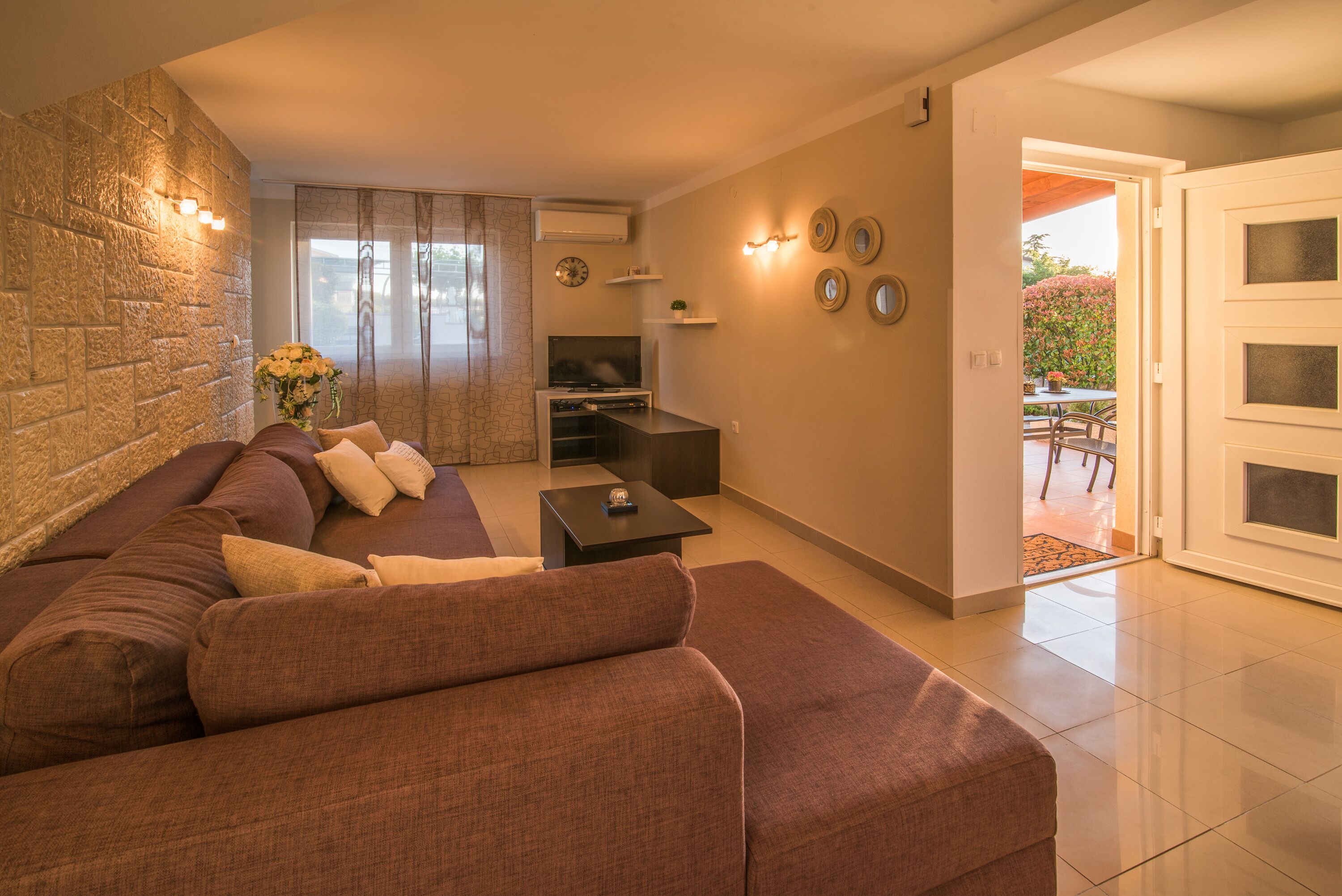 A living room with a brown sofa, a small table, and a TV. There is a stone wall and a door leading to an outdoor area.