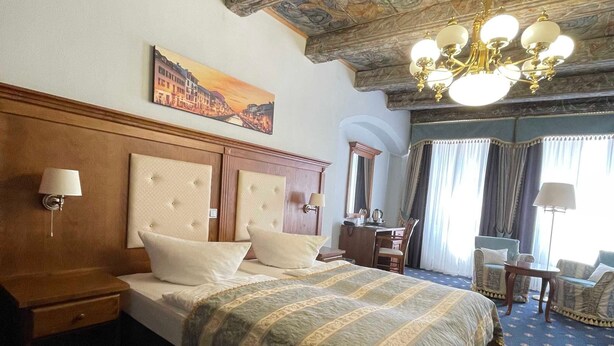 A hotel room with a large bed, a wooden headboard, a chandelier, and a painting of a cityscape. - Double Room Comfort Hotel**** - Hotel Italia (Görlitz)