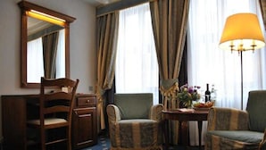 Een hotelkamer met een houten bureau, twee gestoffeerde fauteuils, een tafeltje met een bloemenvaas en een lamp.