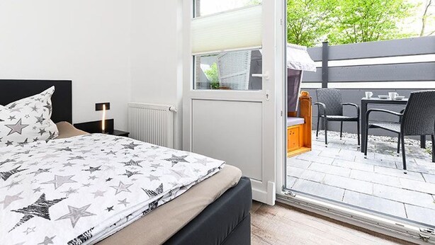 Un dormitorio con cama, una mesita de noche, una ventana y una puerta abierta que da a un patio exterior con mesa y sillas.