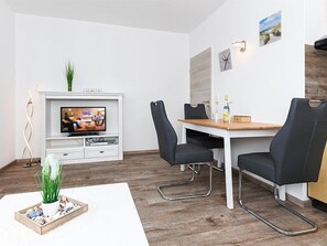 Uma área de jantar moderna com uma mesa de madeira, cadeiras pretas, uma televisão sobre um móvel branco e uma pequena planta no chão.