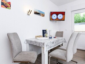 Uma área de refeições com uma mesa posta para dois, duas cadeiras, uma televisão presa na parede e uma janela com persianas.