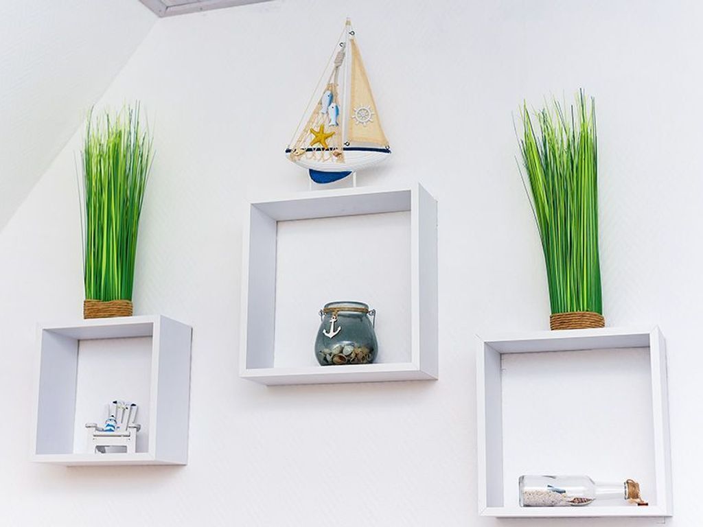 Tres estantes blancos montados en la pared con plantas verdes, un pequeño velero decorativo y un frasco con un ancla.