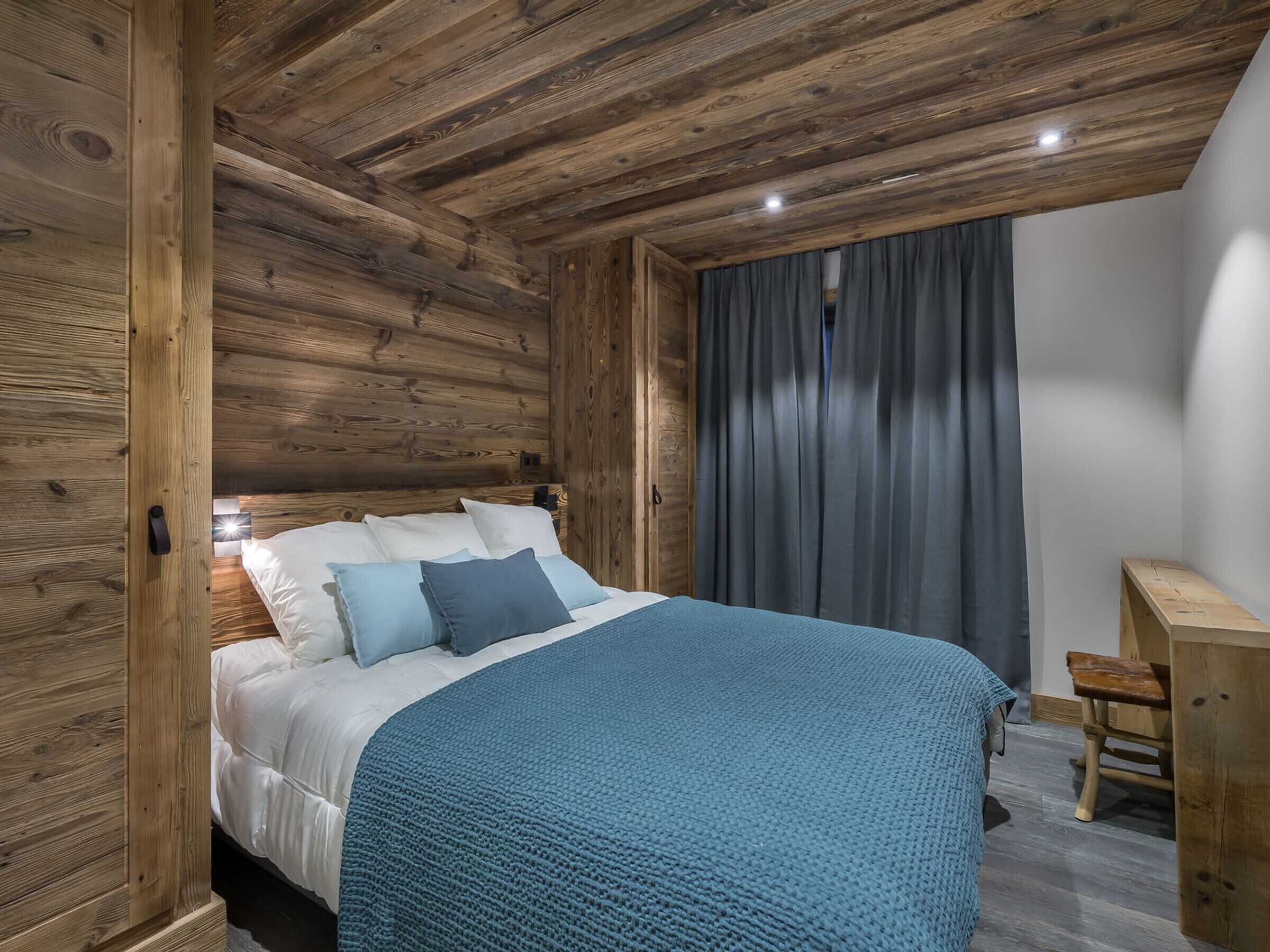A bedroom with a wooden ceiling, a bed with white and blue bedding, a small wooden desk, and a grey curtain.
