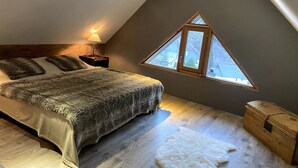 Une chambre avec un plafond en pente, un lit recouvert d’une couverture moelleuse, une table de chevet avec une lampe, une commode en bois et un puits de lumière.