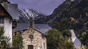 Un bâtiment en pierre doté d’une fenêtre circulaire et d’un clocher, entouré d’arbres et adossé à un paysage montagneux.