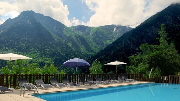 Une piscine entourée d’une clôture en bois, agrémentée de chaises longues et de parasols, avec un paysage montagneux en toile de fond.