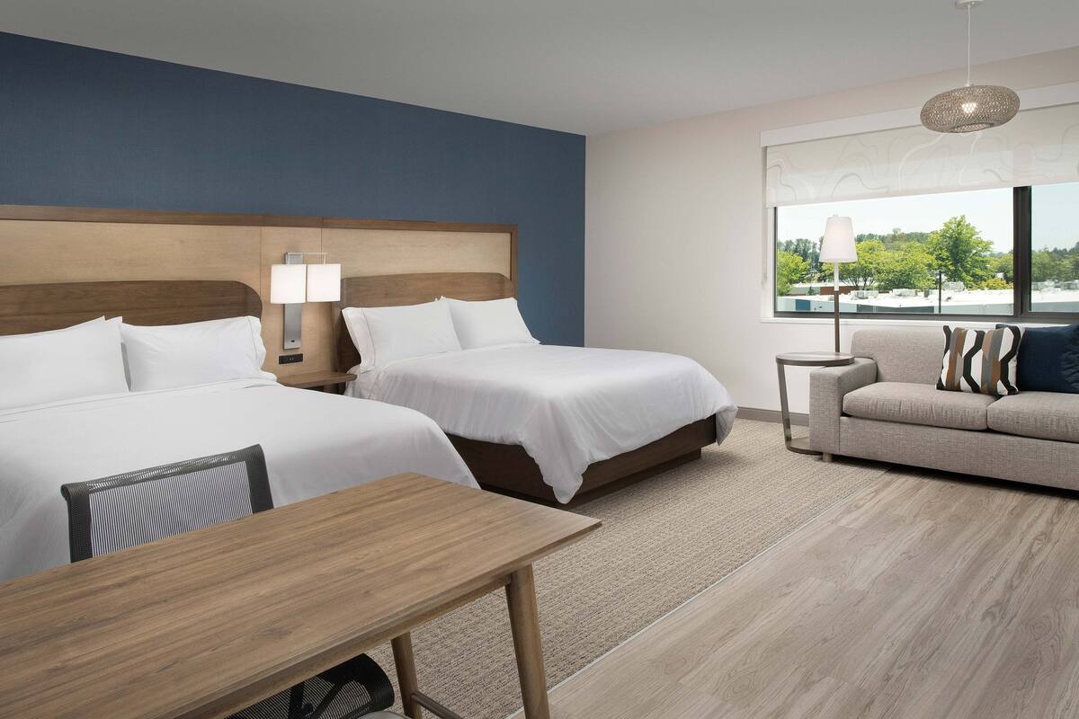 A hotel room with two beds, a sofa, a wooden table, and a large window with a view.