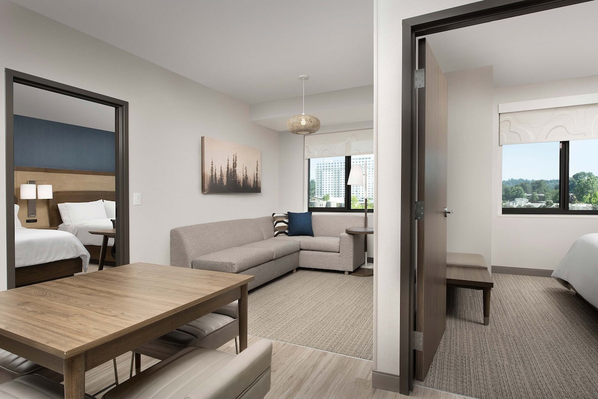 A modern hotel room with a wooden dining table, a sofa, a bed, and a view of the city through the window.