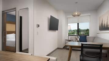 Une chambre d’hôtel moderne avec une table à manger en bois, un fauteuil de bureau noir, un canapé, un téléviseur à écran plat fixé au mur et une fenêtre donnant sur un paysage verdoyant.